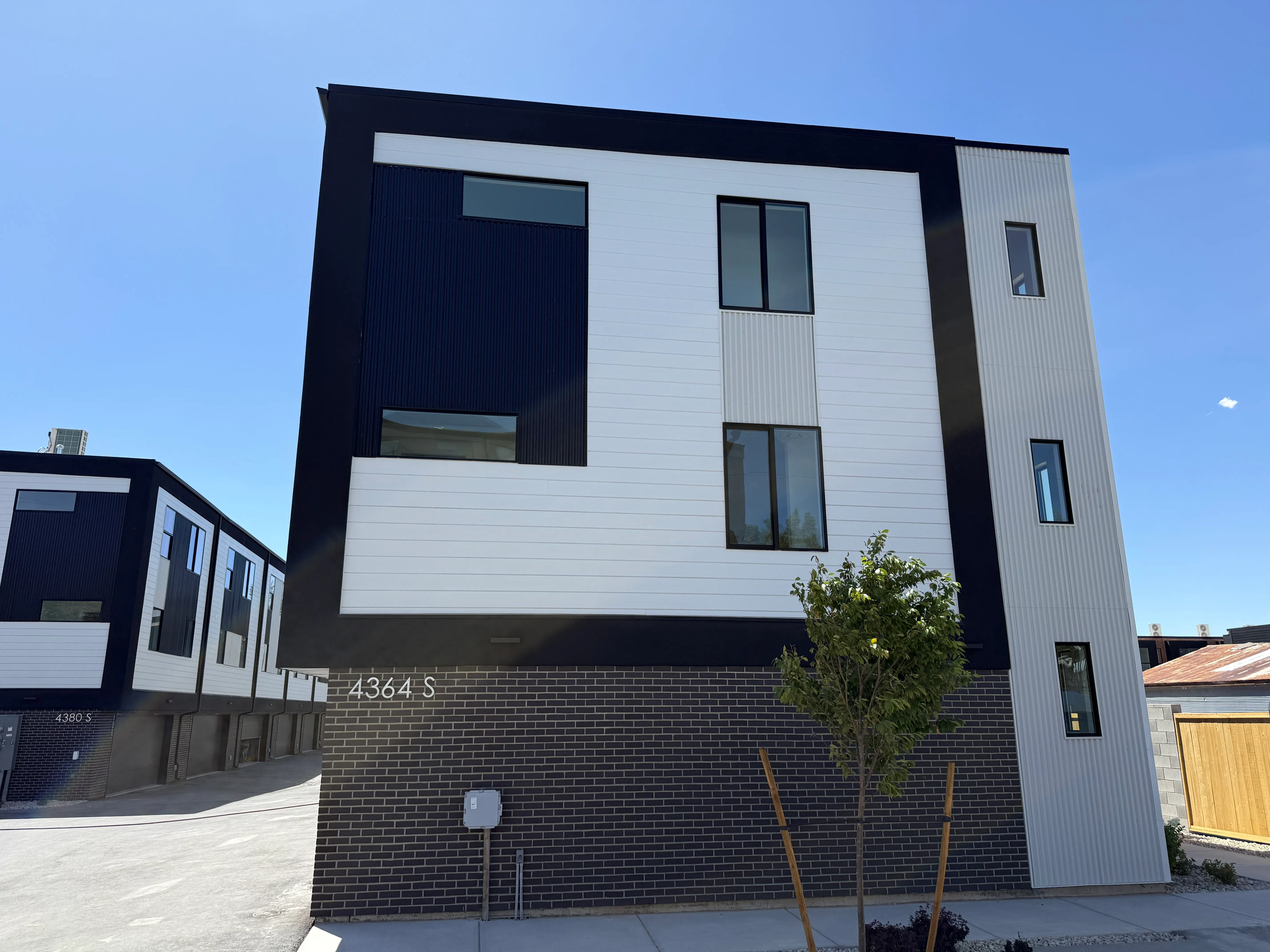 Exterior of a brand-new three-story Quincy townhome with modern black-and-white siding, attached garage, and a young tree against a clear blue sky