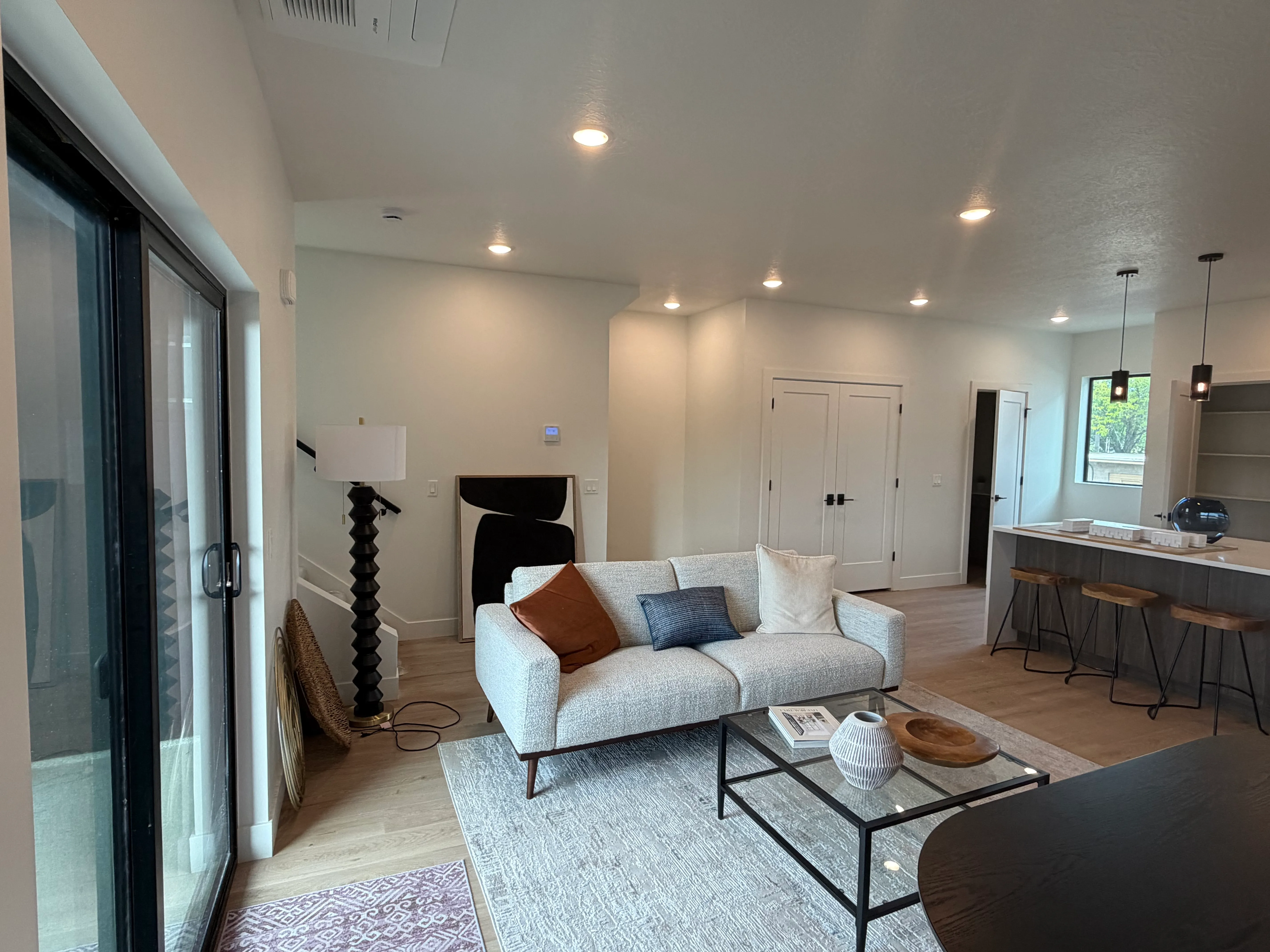 Staged living room with a sofa, glass coffee table, and area rug, open to the kitchen and staircase